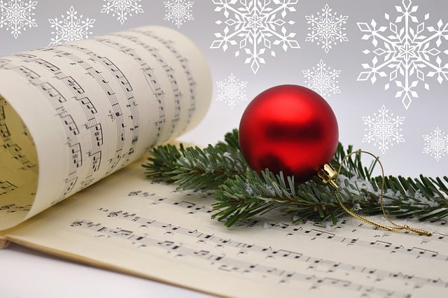 Open book of sheet music, with a pine branch and a Christmas decoration on it. Snowflakes are in the background