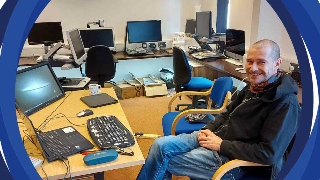 A photo of Duncan in our Resource Centre with a computer and visual aids. The room is filled with many other visual aids in the background.