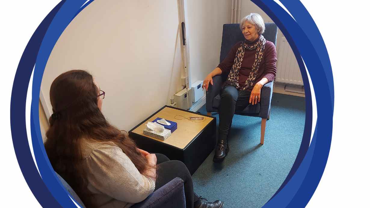 Our counselling room with 2 chairs opposite each other. Our counsellor Jill is sitting at the far chair and is facing the camera. There is a person sitting at the chair closer to the camera, looking away and talking to Jill.