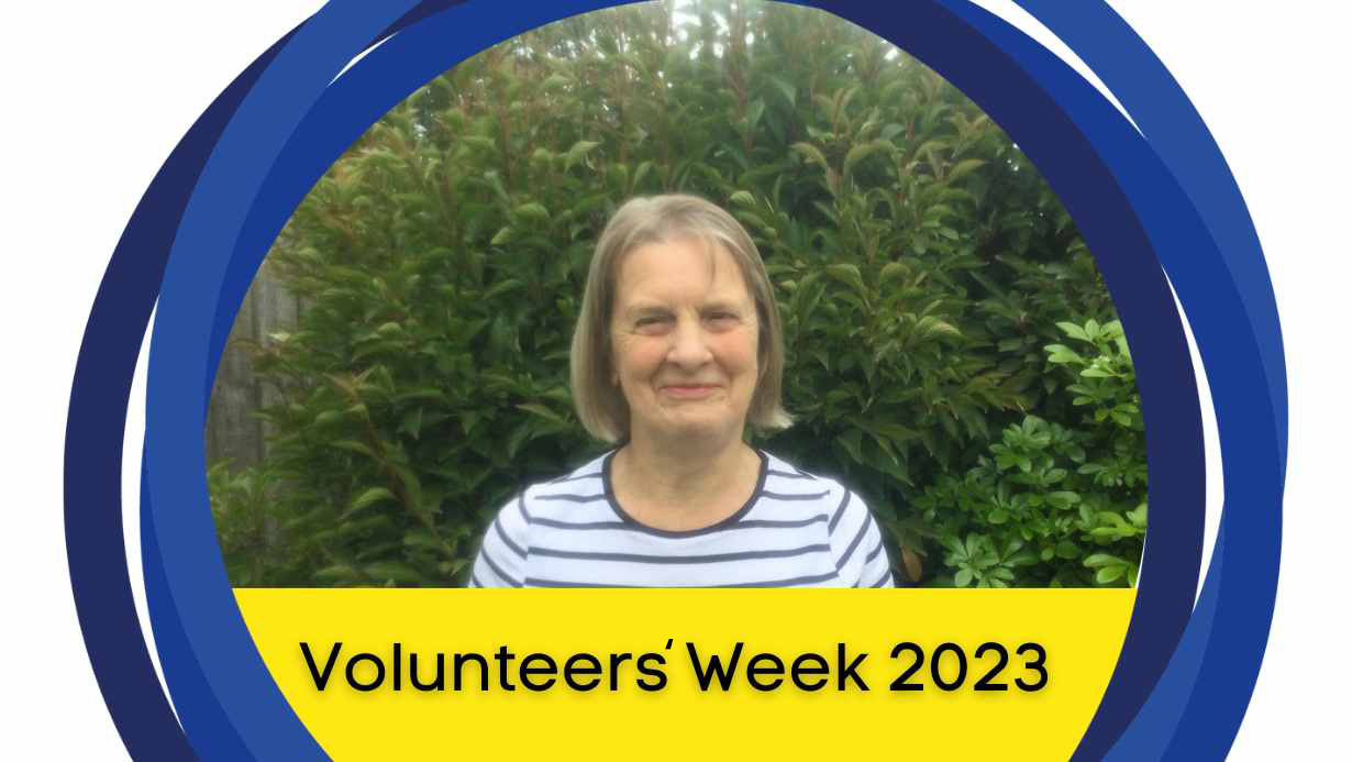 Lesley posing for a photo with green trees and bushes in the background. There is a yellow banner below with text that says, 'Volunteers' Week 2023.'