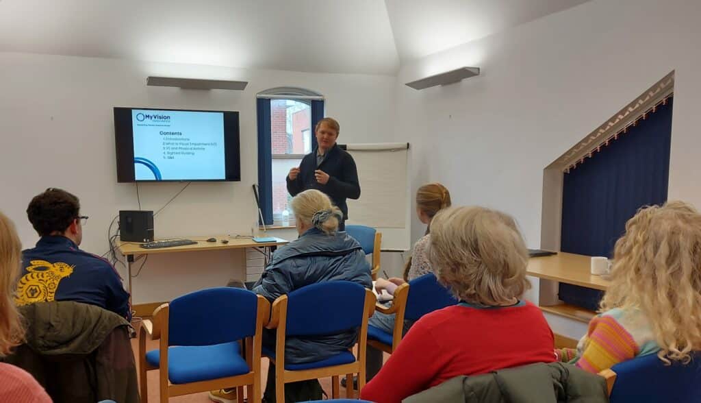 Nathan, our Client Advice Officer giving a Visual Impairment Awareness presentation. 5 people are in the frame, sitting and listening.