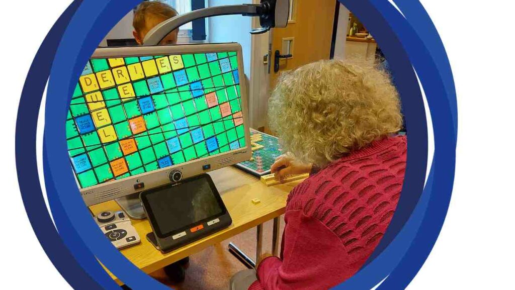 A photo of Jean from behind, using a CCTV magnifier to play Scrabble. The board can be seen on the monitor. 