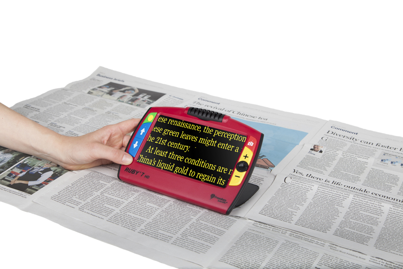 A newspaper with the Ruby 7 on top of it zooming in on part of the text. The text is zoomed in on the magnifier's screen and is in a yellow-on-black colour contrast.