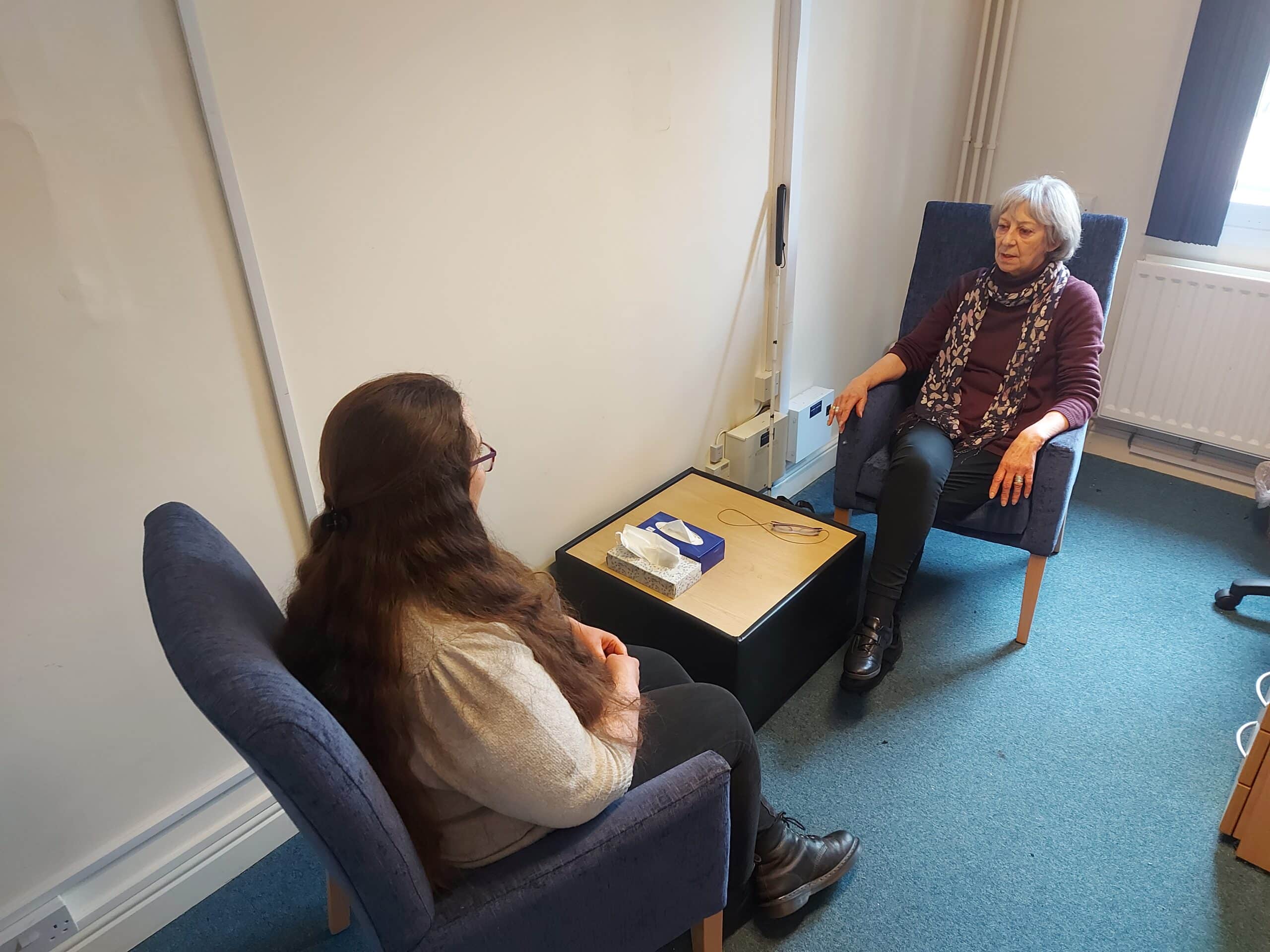 Our counselling room with 2 chairs opposite each other. Our counsellor Jill is sitting at the far chair and is facing the camera. There is a person sitting at the chair closer to the camera, looking away and talking to Jill.