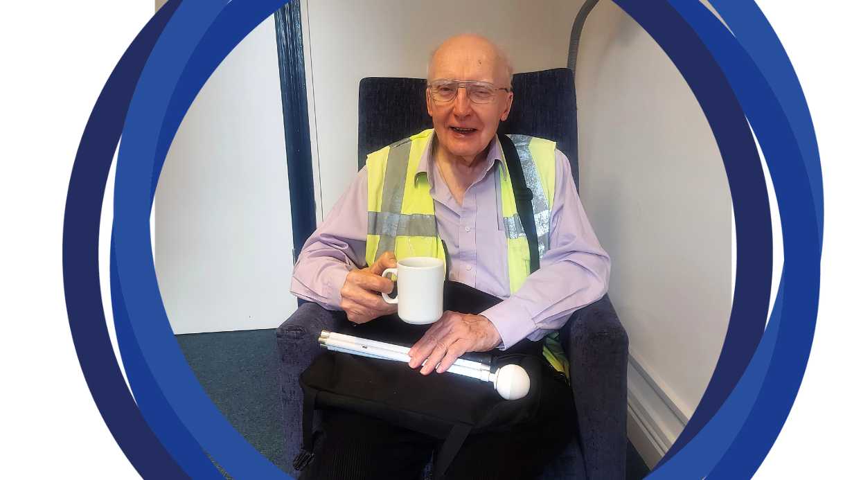 Chris sitting with a cup of tea with his cane on his lap, smiling