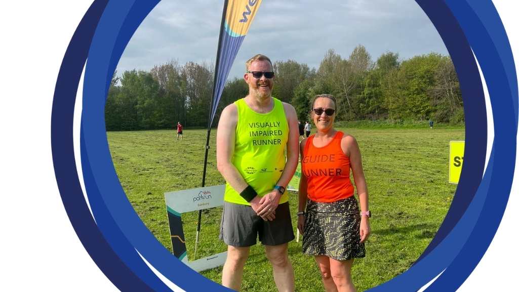 Andy and his guide runner Sharon standing in front of a welcome banner on green grass on a sunny day, smiling and posing in their bright VI runner and Guide Runner tabards